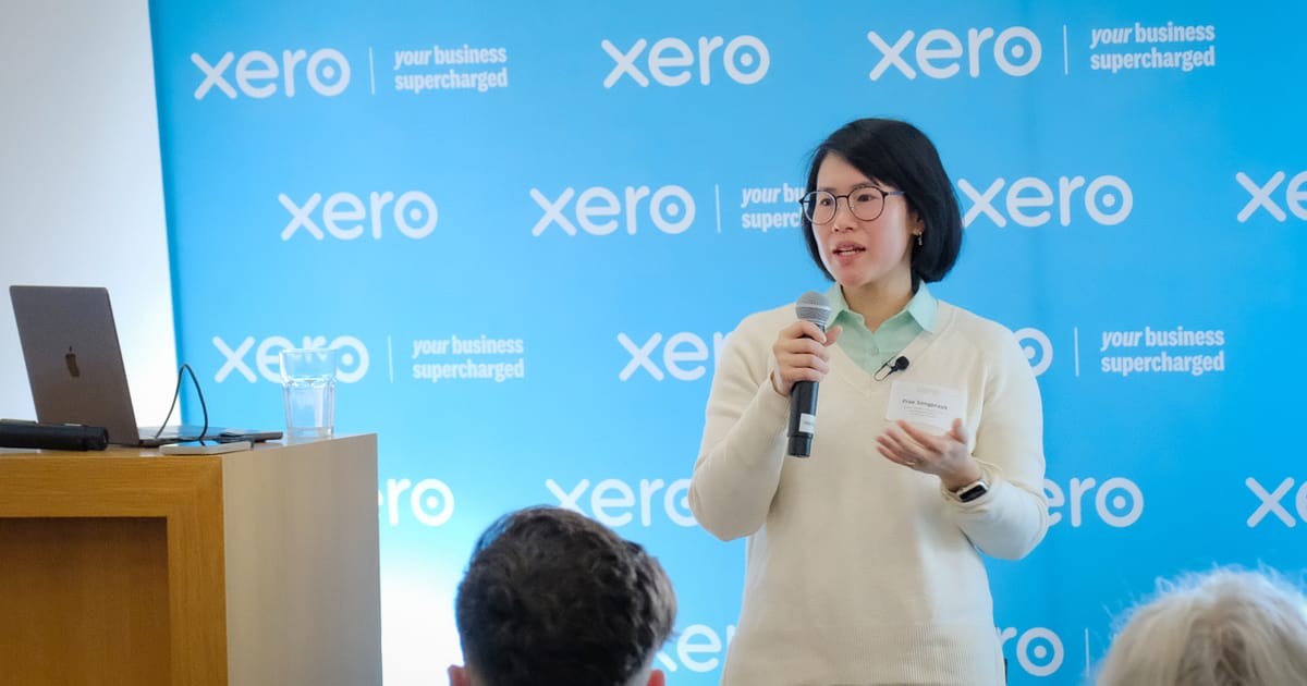 Photo of me, a short haired South East Asian woman with glasses, speaking with a microphone at Code Camp Wellington.